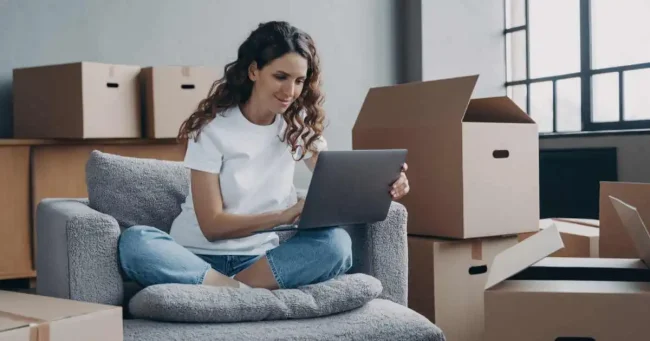 new homeowner on a laptop with moving boxes behind her planning move in cleaning