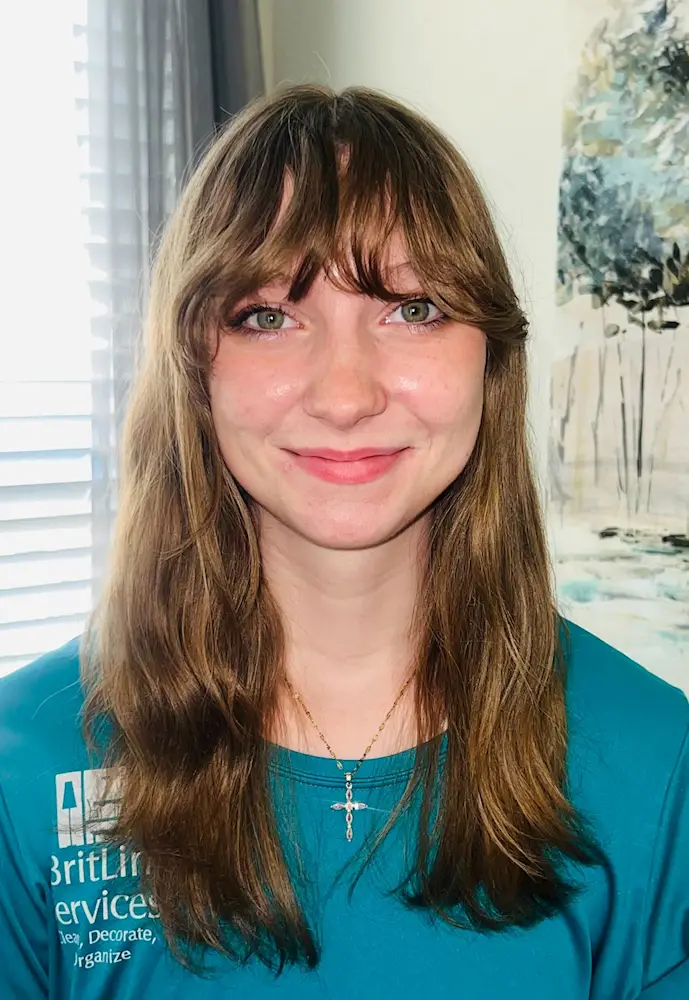 Young woman with long brown hair and bangs smiling at the camera wearing a teal shirt and a cross necklace