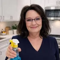 Smiling woman in a navy top holding a spray bottle in a bright kitchen ready to clean a surface