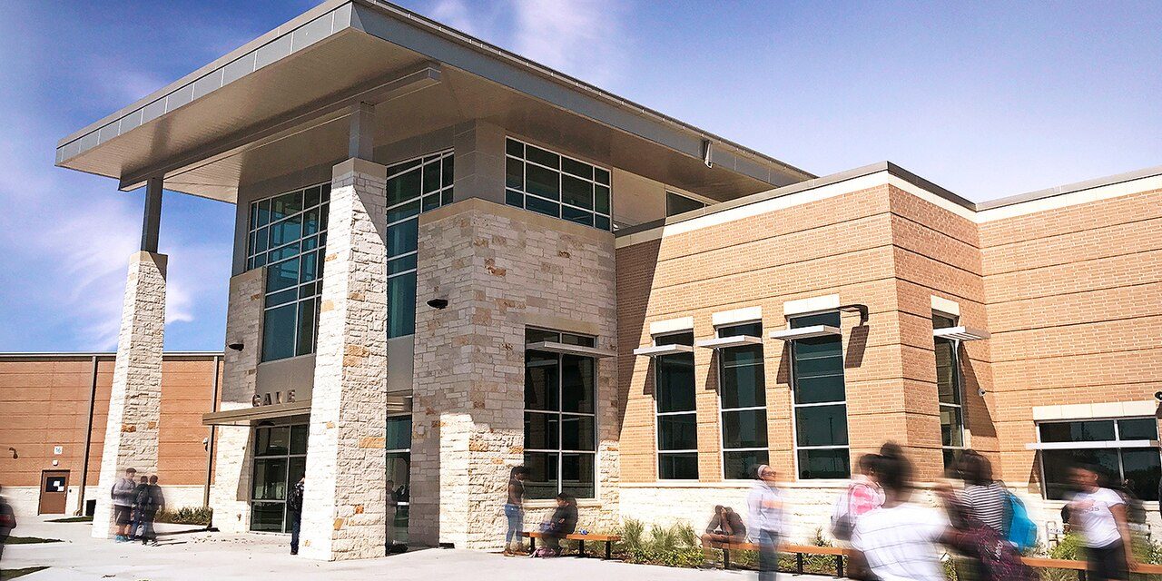 Modern school entrance with stone pillars and brick walls; students walk outside on a sunny day.