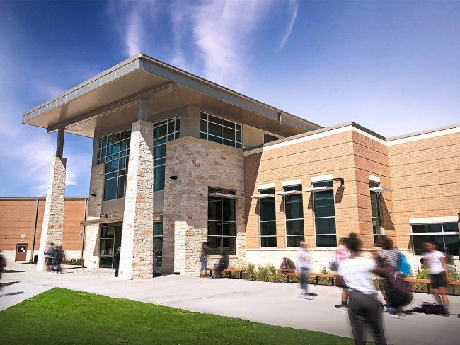 Modern school entrance with stone pillars and brick walls; students walk outside on a sunny day.
